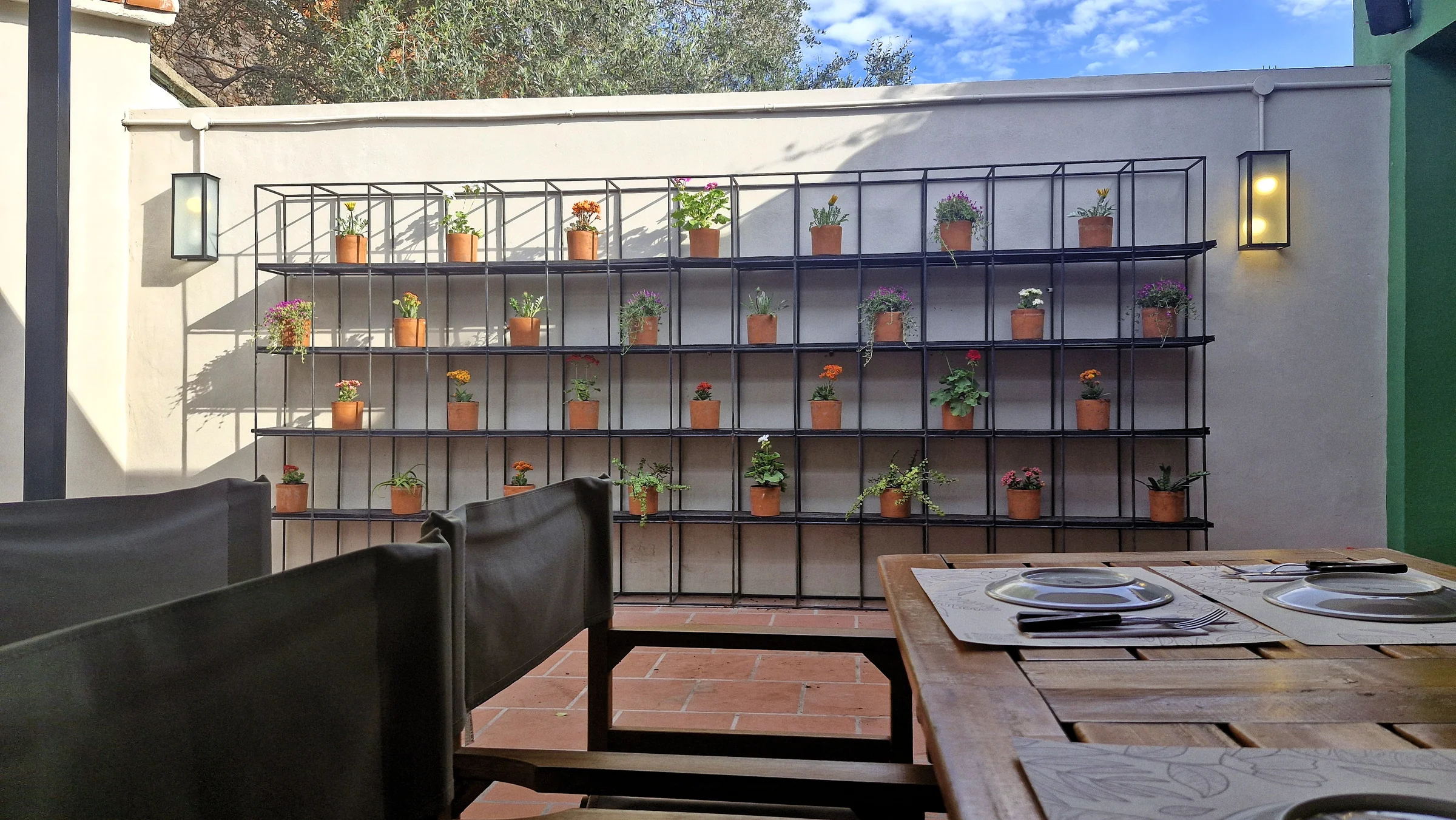 Terraza de Palto: pared de macetas con flores, mesa puesta y apliques encendidos al atardecer